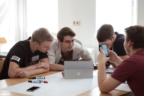 Foto von vier Studenten. Sie sitzen gemeinsam an einem Tisch und schauen auf ein Tablet, das in der Mitte des Tisches steht. 