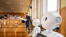 Zwei humanoide Roboter stehen in einem Hörsaal, welcher sich im Hintergrund gerade füllt.