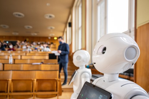 Zwei humanoide Roboter stehen in einem Hörsaal, welcher sich im Hintergrund gerade füllt.