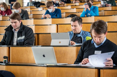 Drei Personen sind in Dokumente oder ihre Laptops vertieft und sitzen in einem Hörsaal.