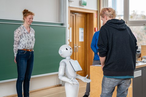 Eine Person redet mit einem humanoiden Roboter, ,eine andere schaut lächelnd zu.