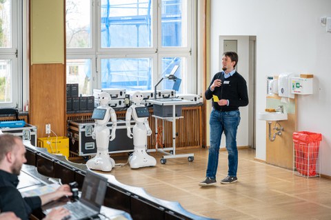 Eine Person hält einen Vortrag, im Hintergrund zwei humanoide Roboter.