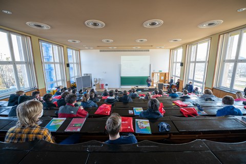 Blick in einen Hörsaal von hinten, viele Menschen, die einem Vortag folgen. 