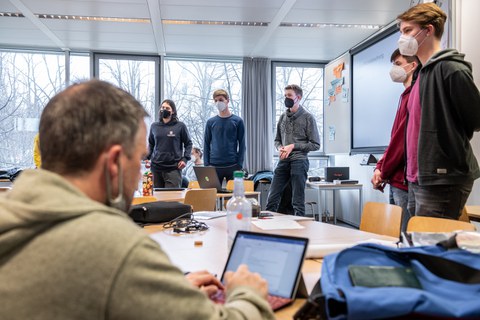 Personen halten einen Vortrag, im Vordergrund macht eine Person mit dem Laptop Notizen.