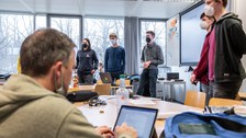 Personen halten einen Vortrag, im Vordergrund macht eine Person mit dem Laptop Notizen.