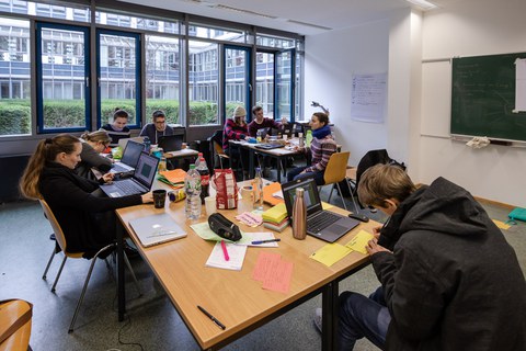 Foto von Studierenden, die in einem Seminarraum verteilt an drei Tischgruppen sitzen und an Laptops arbeiten.