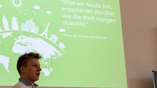 Foto eines Studenten, wie er die Abschlusspräsentation hält. Er hat Notizzettel in den Händen und steht vor einer Wand auf der die Präsentation projiziert wird.