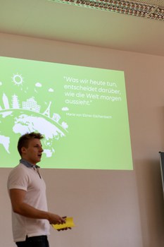Foto eines Studenten, wie er die Abschlusspräsentation hält. Er hat Notizzettel in den Händen und steht vor einer Wand auf der die Präsentation projiziert wird.