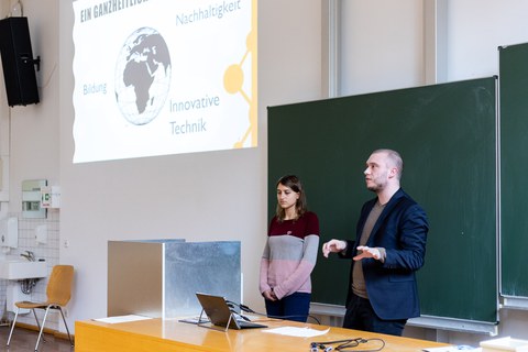 Foto von zwei Studierenden, die vor einer Tafel stehen und präsentieren. Über ihnen wird die Präsentation auf die Wand projiziert.