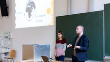 Foto von zwei Studierenden, die vor einer Tafel stehen und präsentieren. Über ihnen wird die Präsentation auf die Wand projiziert.