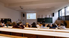 Foto eines Hörsaales von hinten betrachtet. Viele Studierende sitzen in den Sitzreihen und schauen nach vorne. Dort wird an die Wand eine Präsentation projiziert.