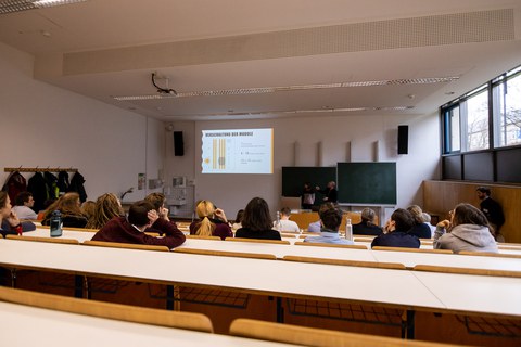 Foto eines Hörsaales von hinten betrachtet. Viele Studierende sitzen in den Sitzreihen und schauen nach vorne. Dort wird an die Wand eine Präsentation projiziert.