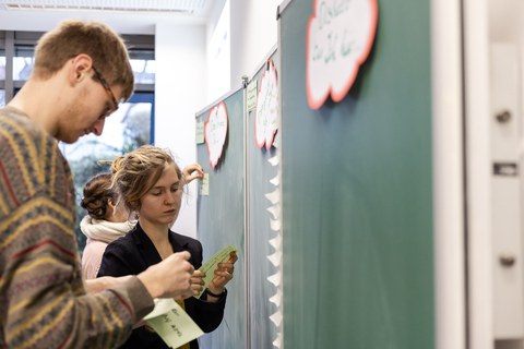 Foto von der Methode zum Abgleich der Erwartungshaltung. Studierende stehen vor einer Tafel und bekleben diese mit beschrifteten Zetteln.