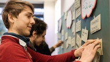 Foto von der Methode zum Abgleich der Erwartungshaltung. Zwei Studenten stehen vor einer Tafel und bekleben diese mit beschrifteten Zetteln.