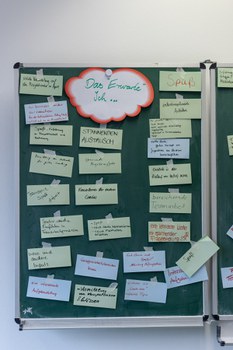 Foto von dem Ergebnis der Methode über die Erwartungshaltung. An einer Tafel klebt oben in der Mitte ein Zettel in wolkenform mit dem Text: "Das erwarte ich". Daneben und darunter befinden sich weitere beschriftete Zettel mit den Antworten der Coaches.