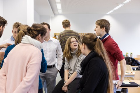 Foto von den Coaches. Sie sitzen oder stehen in einem Seminarraum und unterhalten sich.