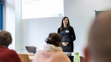 Foto von der Vorstellung der Aufgabenstellung. Eine Frau steht neben einer Tafel und erklärt etwas.