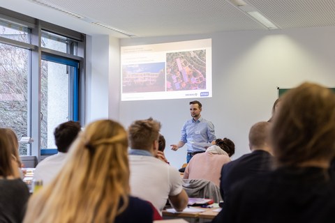 Foto von der Vorstellung der Aufgabenstellung. Ein Mann steht neben einer Tafel und erklärt etwas. Vor ihm sitzen Studierende in Reihen an Tischen und hören ihm zu.
