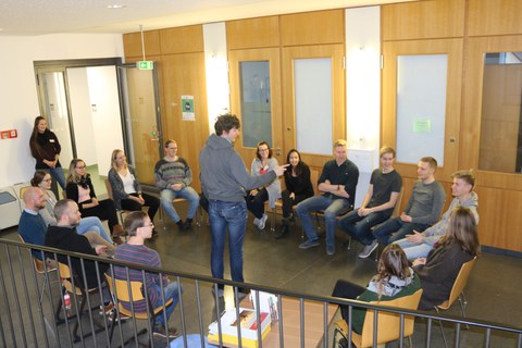 Foto während einer Kennenlernmethode. Die Studierenden sitzen in einem Stuhlkreis und ein Student steht in der Mitte des Kreises und spricht.