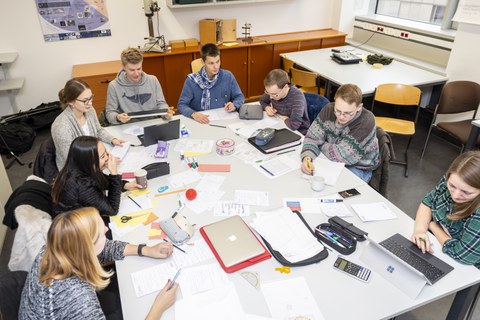 Foto von Studierenden während des Arbeitsprozesses. Die Studierenden sitzen um einen großen Tisch und diskutieren miteinander oder arbeiten an ihren Laptops. Der Tisch ist von Arbeitsmaterialien wie Blöcken, Stiften und Taschenrechnern bedeckt.