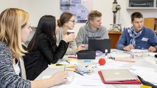 Foto von Studierenden, die an einem großen Tisch sitzen und auf Blöcken schreiben oder an ihren Laptops arbeiten.