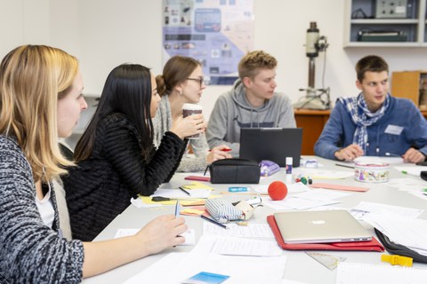 Foto von Studierenden, die an einem großen Tisch sitzen und auf Blöcken schreiben oder an ihren Laptops arbeiten.