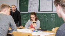 Foto einer Studentin, die an einem Pult vor einer voll beschriebenen Tafel sitzt. Sie schaut vor sich auf den Tisch auf dem ein beschriebenes Flipchartpapier liegt. Drei weitere Studenten stehen oder sitzen um das Pult und betrachten auch das Papier.