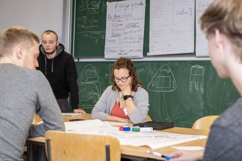 Foto einer Studentin, die an einem Pult vor einer voll beschriebenen Tafel sitzt. Sie schaut vor sich auf den Tisch auf dem ein beschriebenes Flipchartpapier liegt. Drei weitere Studenten stehen oder sitzen um das Pult und betrachten auch das Papier.