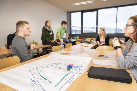 Foto einer Gruppe Studierender, die um einen großen Tisch sitzt und miteinander diskutiert. Auf dem Tisch liegt ein beschriftetes Flipchartpapier sowie Stifte.