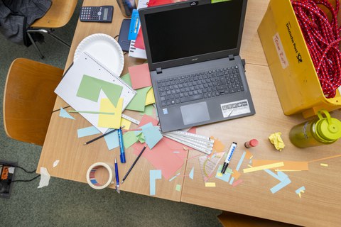 Foto eines Tisches von oben gesehen. Auf dem Tisch steht ein aufgeklappter Laptop und es liegen viele Arbeitsmaterialien wie bunte Blätter, Stifte, Kreppband, Lineale, Kleber sowie ein Taschenrechner daneben.