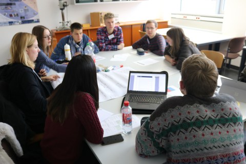 Foto einer Gruppe Studierender, die um einen großen Tisch sitzt und miteinander diskutiert. Auf dem Tisch liegt ein beschriftetes Flipchartpapier sowie Stifte und weitere Blöcke und ein aufgeklappter Laptop.