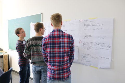 Foto von drei Studenten, die drei beschriftete Flipchartpapiere betrachten, die vor ihnen an der Wand hängen.
