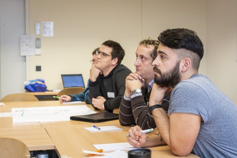 Foto der Profirunde. Die Profis sitzen in einer Reihe an einem langen Tisch und hören jemandem zu, der sich außerhalb des Fotos befindet.