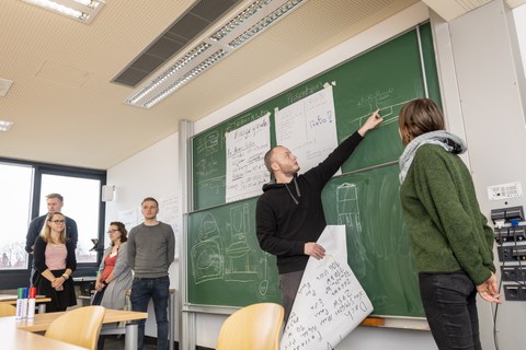 Foto einer Gruppe Studierender, die vor einer beschrifteten Tafel steht. Ein Student zeigt auf die Tafel und erklärt etwas.