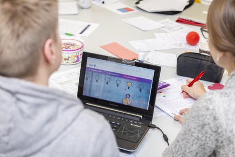Foto von zwei Studierenden, die an einem Tisch sitzen und arbeiten. Ein Student arbeitet an einem Laptop und die Studentin rechts neben ihm schreibt mit einem Stift auf einem Block.