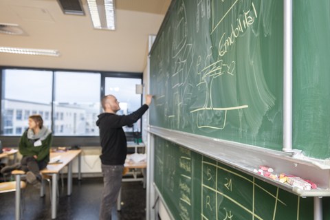 Foto eines Studenten, der vor einer Tafel steht und diese mit Kreide beschriftet.