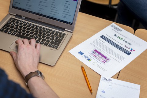 Foto eines Tisches von schräg oben betrachtet. Auf ihm liegen ein Block, ein Stift und ein Zettel mit BeING Inside Schriftzug. Weiter steht ein aufgeklappter Laptop auf dem Tisch und man erkennt eine Hand, die auf dem Laptop das Touchpad bedient.