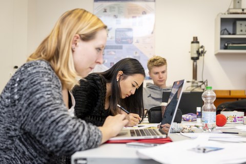 Foto von drei Studierenden, die an einem Tisch sitzen und an ihren Laptops arbeiten.