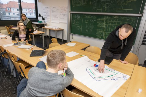 Foto eines Seminarraumes, in dem sich vier Studierende befinden. Zwei Studenten stehen sich an einem Tisch gegenüber und betrachten ein beschriftetes Flipchartpapier, das vor ihnen auf dem Tisch liegt.
