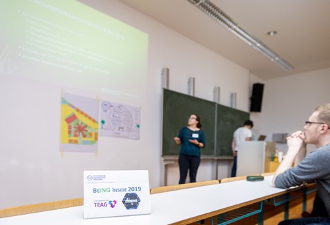 Foto eines Hörsaales. Vorne vor der Tafel steht eine Frau und erklärt etwas. Eine Präsentation wird links über ihr an die Wand projiziert. 