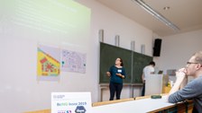Foto eines Hörsaales. Vorne vor der Tafel steht eine Frau und erklärt etwas. Eine Präsentation wird links über ihr an die Wand projiziert. 