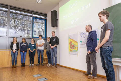 Foto einer Studierendengruppe bei der Abschlusspräsentation. Die Studierenden stehen in einer Reihe vor einer Wand. Über ihnen wird eine Präsentation an die Wand projiziert. Neben ihnen an der Wand hängen zwei beschriftete Flipchartpapiere.
