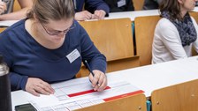 Foto einer Frau, die in einer Sitzreihe in einem Hörsaal sitzt und einen Bewertungsbogen ausfüllt.