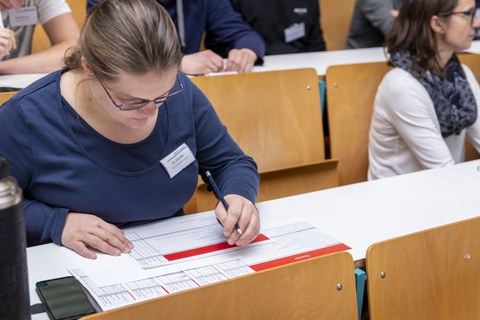 Foto einer Frau, die in einer Sitzreihe in einem Hörsaal sitzt und einen Bewertungsbogen ausfüllt.