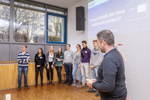 Foto einer Studierendengruppe bei der Abschlusspräsentation. Die Studierenden stehen in einer Reihe vor einer Wand. Über ihnen wird eine Präsentation an die Wand projiziert mit dem Text: "Danke für Ihre Aufmerksamkeit".