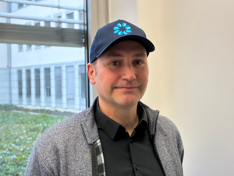 Ein Mann mit dunkelblauem Basecap steht lächelnd vor einem Fenster.