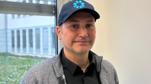 Ein Mann mit dunkelblauem Basecap steht lächelnd vor einem Fenster.