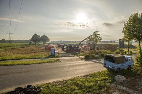 Im Vordergrund eine Landstraße mt einem kleinen Lieferwagen. Im Hintergrund ein Feld mit einem Baukran auf einer Baustelle.