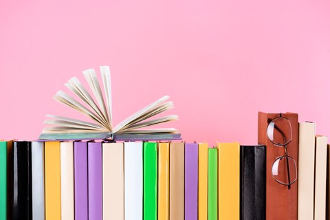  Das Foto zeigt eine Reihe von Büchern vor einem rosa Hintergrund. Ein Buch liegt aufgeschlagen auf den anderen. An einem anderen Buch hängt eine Brille.