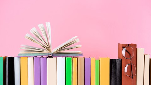  Das Foto zeigt eine Reihe von Büchern vor einem rosa Hintergrund. Ein Buch liegt aufgeschlagen auf den anderen. An einem anderen Buch hängt eine Brille.